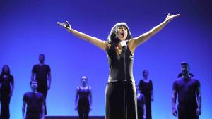 Foto de uma cena do espetáculo que mostra a atriz cantando de frente para um microfone, com os braços abertos para o alto. Atrás, bailarinos com roupas pretas fazem coro.