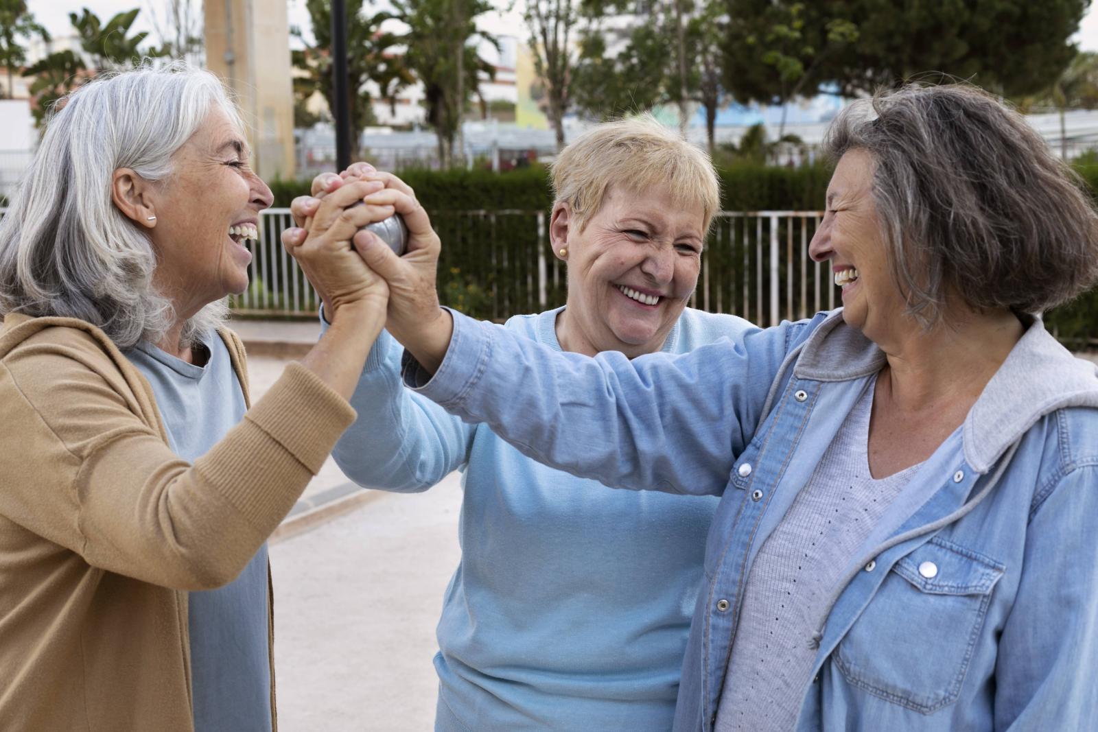 Três idosas rindo em momento de lazer, em uma área aberta.